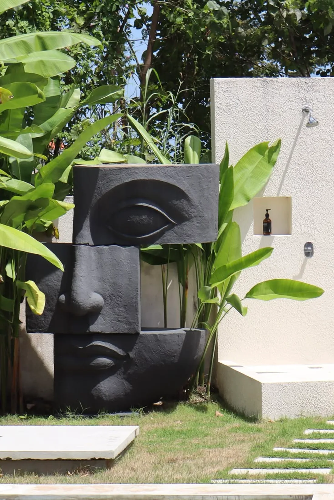 Escultura de um rosto monumental montado em blocos escuros, com olho, nariz e boca, em meio a plantas de folhas grandes